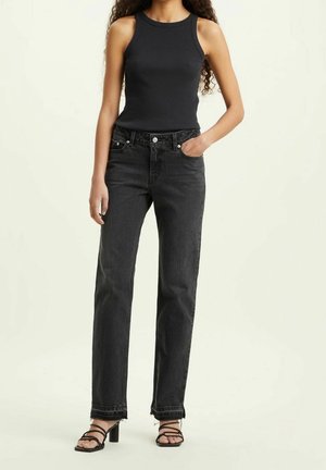 Persoon draagt een mouwloos zwart topje, hoge taille zwarte spijkerbroek met rafelige zomen en zwarte sandalen met hak en bandjes, staand met één hand in de zak.