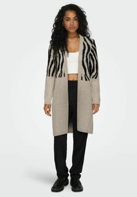 Beige lange cardigan met zwarte zebra-stripes, gemaakt van zachte garen, met open voorkant en ribbelmanchetten, gedragen over een witte top en zwarte broek.
