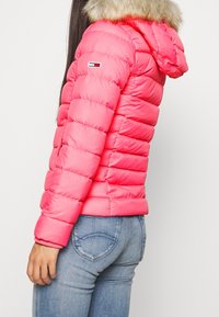 Růžová péřová bunda s kapucí podšitou kožešinou, s prošívanými částmi a logem na rukávu; v kombinaci s modrými džíny.