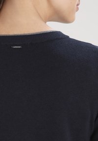 Marinblå tröja med en ribbad halsringning och grå accent, slät textur och en liten metallisk logotyp baktill.