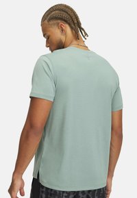 Camiseta verde de manga corta con un patrón texturizado, que presenta una abertura lateral y un escote redondo, llevada por una persona vista desde atrás.