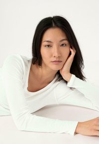 Top bianco a maniche lunghe indossato da un modello, con scollatura rotonda e tessuto morbido e strutturato, posato su uno sfondo neutro.