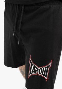 Juodi medvilniniai šortai su elastinga juosta ir raišteliu. Savybė - ryškus "TAPOUT" logotipas raudonomis ir baltomis spalvomis kairėje kojoje.