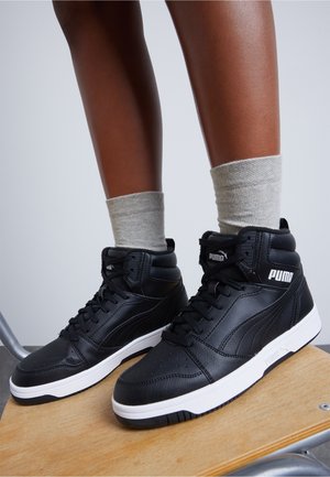 Person, die schwarze Puma High-Top-Sneaker mit weißen Sohlen und hellgrauen Socken trägt und auf einem Holzstuhl vor einem schlichten Hintergrund steht.