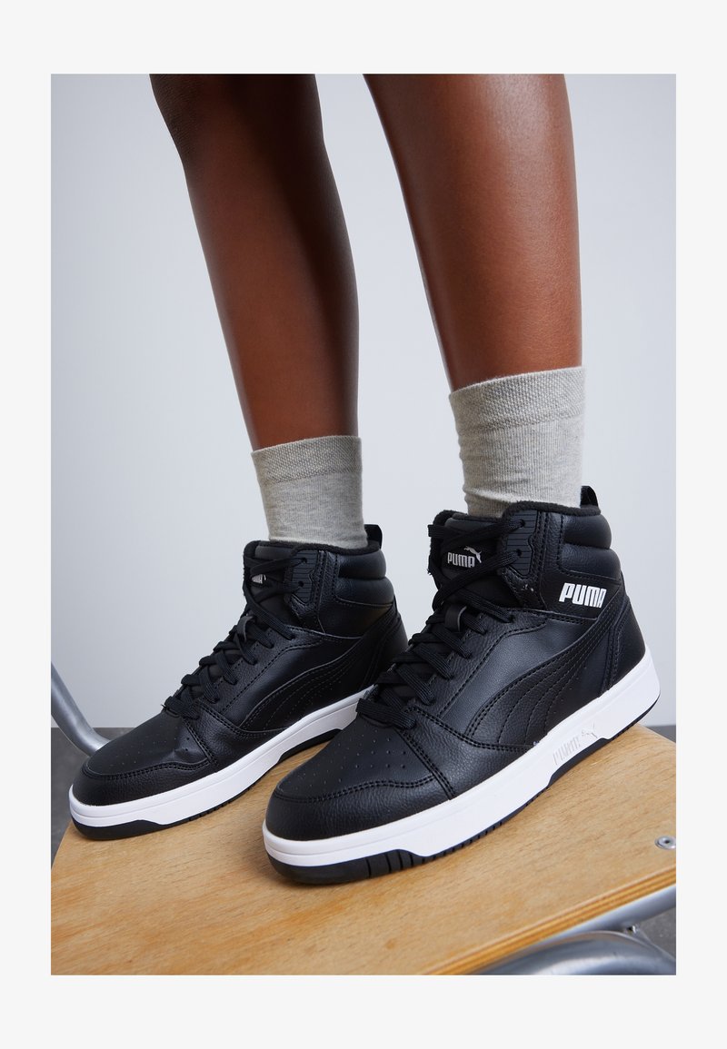 Personne portant des baskets montantes noires Puma avec des semelles blanches et des chaussettes gris clair, debout sur une chaise en bois devant un fond uni.