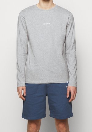 Personne portant une chemise à manches longues gris clair avec le texte "LES DEUX" et un short bleu marine, debout devant un fond uni clair.