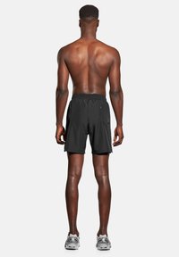 Czarne sportowe szorty z elastycznym pasem, z pojedynczą boczną kieszenią, noszone z szarymi sneakersami. Widok z tyłu.