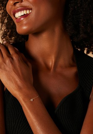 Gouden armband met een kleine decoratieve accent, gedragen op een donkerhuidige arm, met een textuur zwart top zichtbaar.