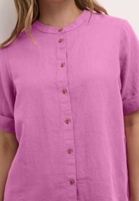 Kortärmad, rosa linneskjorta med mandarin-krage, som har sex bruna knappar framtill och en avslappnad passform.
