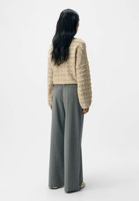 Beige gestrickter Cropped-Pullover mit strukturierten Mustern, kombiniert mit grauen, gestreiften, weiten Hosen. Schuhe in neutralen Farbtönen runden das Outfit ab.