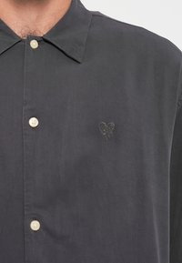 Cămașă button-up de culoare gri închis, confecționată dintr-un material moale, având o guler ascuțit, nasturi deschiși la culoare și un mic detaliu brodat sub formă de inimă pe piept.