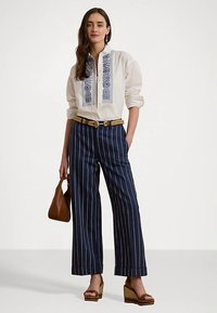 Camisa branca com detalhe bordado azul, combinada com calças largas às riscas azul-marinho e brancas, cinto castanho e sandálias de cunha.