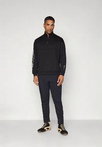 Must kapuutsiga lukuga dressipluus pikkade varrukatega, mille ülaosa on ühevärviline ja vasakul käel on logoga detail. Kombineeritud tumedate kitsaste pükste ja jalanõudega.