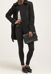 Nainen, jolla on päällään musta takki, mustat housut vetoketjullisilla taskuilla, valkoinen paita, mustat tupsulöysi ja hän pitää mustaa clutch-laukkua.