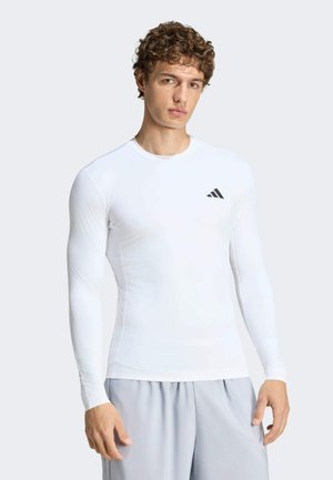 Mężczyzna ubrany w dopasowaną białą sportową koszulkę z długim rękawem z małym czarnym logo Adidas na piersi i jasnoszare szorty.