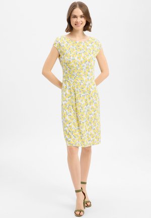 Junge Frau steht mit den Händen hinter dem Rücken, trägt ein gelbes, kurzärmeliges Kleid mit Zitronenmuster in Knie-Länge und grüne Schuhe mit Knöchelriemen.