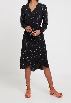 Czarna sukienka z długim rękawem i dekoltem w serek, ozdobiona białym abstrakcyjnym wzorem. Sukienka ma talię podkreśloną paskiem oraz asymetryczny dół, w komplecie z pomarańczowymi sandałami na paski.