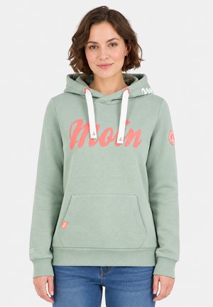 Vrouw draagt lichtgroene hoodie met witte trekkoorden en koraalkleurige "Moin" tekst, gecombineerd met blauwe spijkerbroek, staand tegen een witte achtergrond.