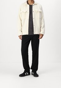 Kurtka polarowa w kolorze ecru z dwoma kieszeniami na piersi, zapinana na zatrzaski, noszona na ciemnym swetrze o fakturze, zestawiona z czarnymi spodniami i trampkami.