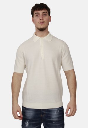 Giovane uomo con capelli ricci e scuri che indossa una polo a maniche corte color avorio e jeans blu scuro strappati, in piedi davanti a uno sfondo neutro.