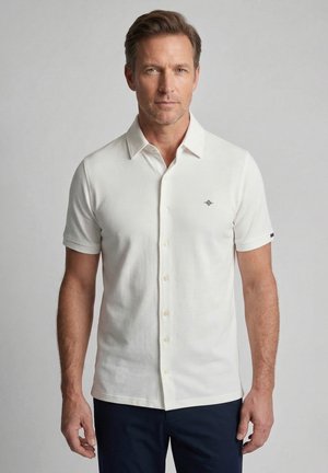 Man draagt een wit kortemouwen poloshirt met klein borstlogo en donkere broek, staand tegen een effen lichtgrijze achtergrond.