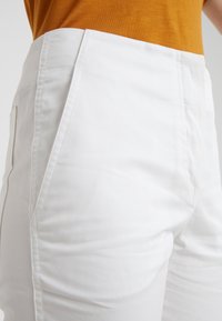 Bílé kalhoty s hladkým bavlněným povrchem, se bočními kapsami a přizpůsobenými švy. Pasový lem kontrastuje s hořčicovým vrškem.