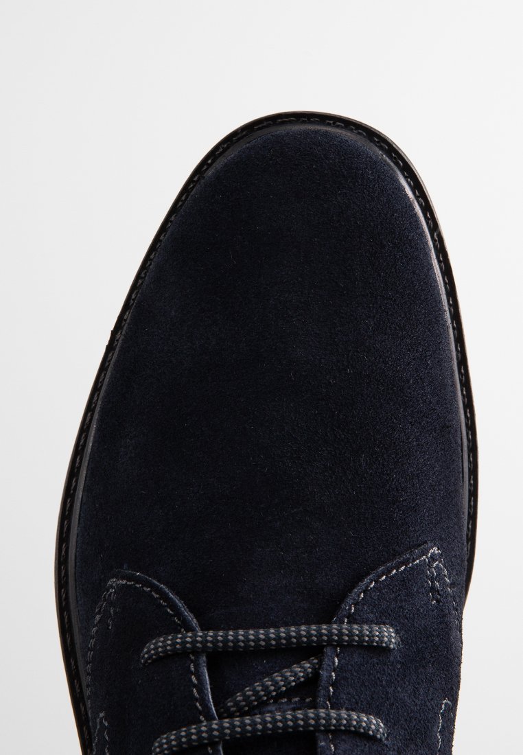Close-up van de punt van een donkerblauwe suede schoen met gestikte randen en een gespannen voorkant op een effen witte achtergrond.