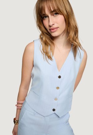 Donna con capelli castano chiaro che indossa un gilet azzurro chiaro senza maniche con bottoni e pantaloni abbinati su uno sfondo uniforme.