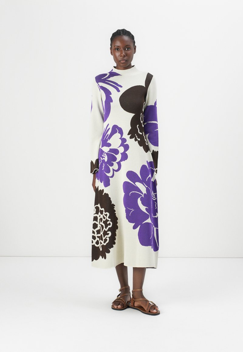 Knee-length knit dress in cream, featuring large purple and brown floral patterns, with long sleeves and a high neckline. Brown sandals.