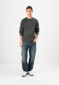 Gray knitted sweater, blue denim jeans, and white sneakers with gray accents. Model stands straight with hands in pockets against a white background.