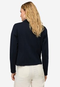 Chaqueta entallada de color azul marino con mangas largas, tejido texturizado, costuras en contraste y un cuello clásico. La vista trasera muestra una silueta estructurada.