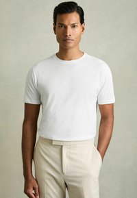 Tricou alb cu mânecă scurtă, cu guler rotund, textură netedă și croială lejeră, combinat cu pantaloni bej deschis.