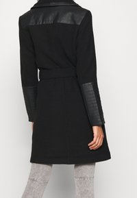 Abrigo negro con cinturón en la cintura, detalles en cuero texturizado en las mangas y los hombros, y un diseño ajustado que llega hasta la mitad del muslo.