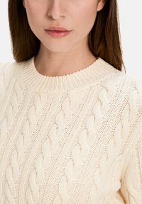 Light cream sweater featuring a textured cable knit pattern, a ribbed neckline, and thick knit construction.
