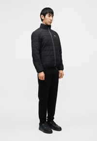 Μαύρο παλτό με καπιτονέ ύφασμα και ψηλό γιακά, κλείνει με φερμουάρ, συνδυασμένο με μαύρο παντελόνι φόρμας και μαύρα αθλητικά παπούτσια. Απλός, μίνιμαλ σχεδιασμός.