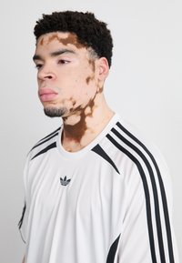 White athletic t-shirt featuring black three-stripe detailing on the shoulders and Adidas logo on the chest. Smooth texture, casual fit.