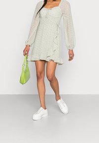 Vestido floral verde claro con mangas largas de tul, dobladillo con volantes y detalle de cinturón anudado. Combinado con zapatillas de plataforma blancas y un bolso verde.