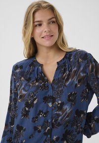 Donna bionda che sorride leggermente, indossando una camicetta blu con motivo floreale e maniche lunghe, che guarda di lato contro uno sfondo semplice.