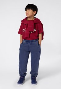 Jongen in een rood Champion t-shirt met een bijpassende sweatshirt over zijn schouders, blauwe joggingbroek en blauwe sneakers, staand met zijn handen in zijn zakken.