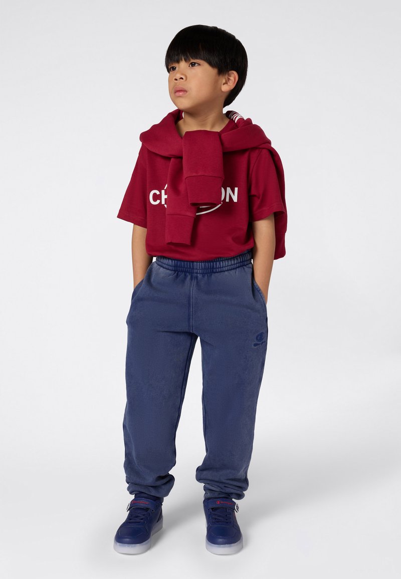 Jongen in een rood Champion t-shirt met een bijpassende sweatshirt over zijn schouders, blauwe joggingbroek en blauwe sneakers, staand met zijn handen in zijn zakken.