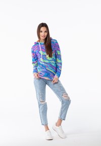 Sudadera con capucha con un patrón de mármol multicolor en azul, rosa, verde y amarillo. Combinada con unos jeans claros rasgados y zapatillas blancas.