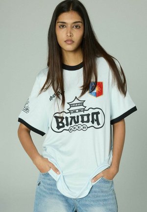 Junge Frau mit langem, dunklem Haar trägt ein weißes Binda Beer T-Shirt mit schwarzem Rand und hellblauen Jeans, Hände in den Taschen, neutraler grauer Hintergrund.