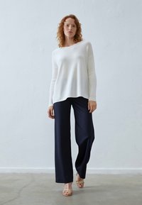 Femeie cu părul creț, îmbrăcată cu un pulover alb cu mâneci lungi, pantaloni largi bleumarin și sandale bej cu bretele, pozând în fața unei pereți albe.