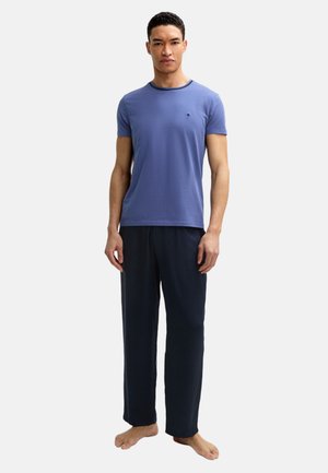 Junger Mann, barfuß stehend, trägt ein blaues T-Shirt mit kurzen Ärmeln und dunkle, locker sitzende Hosen vor einem schlichten hellen Hintergrund.