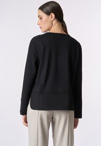 Top nero a maniche lunghe realizzato in tessuto liscio, con un girocollo e un retro leggermente più lungo. Abbinato a pantaloni grigi chiari.