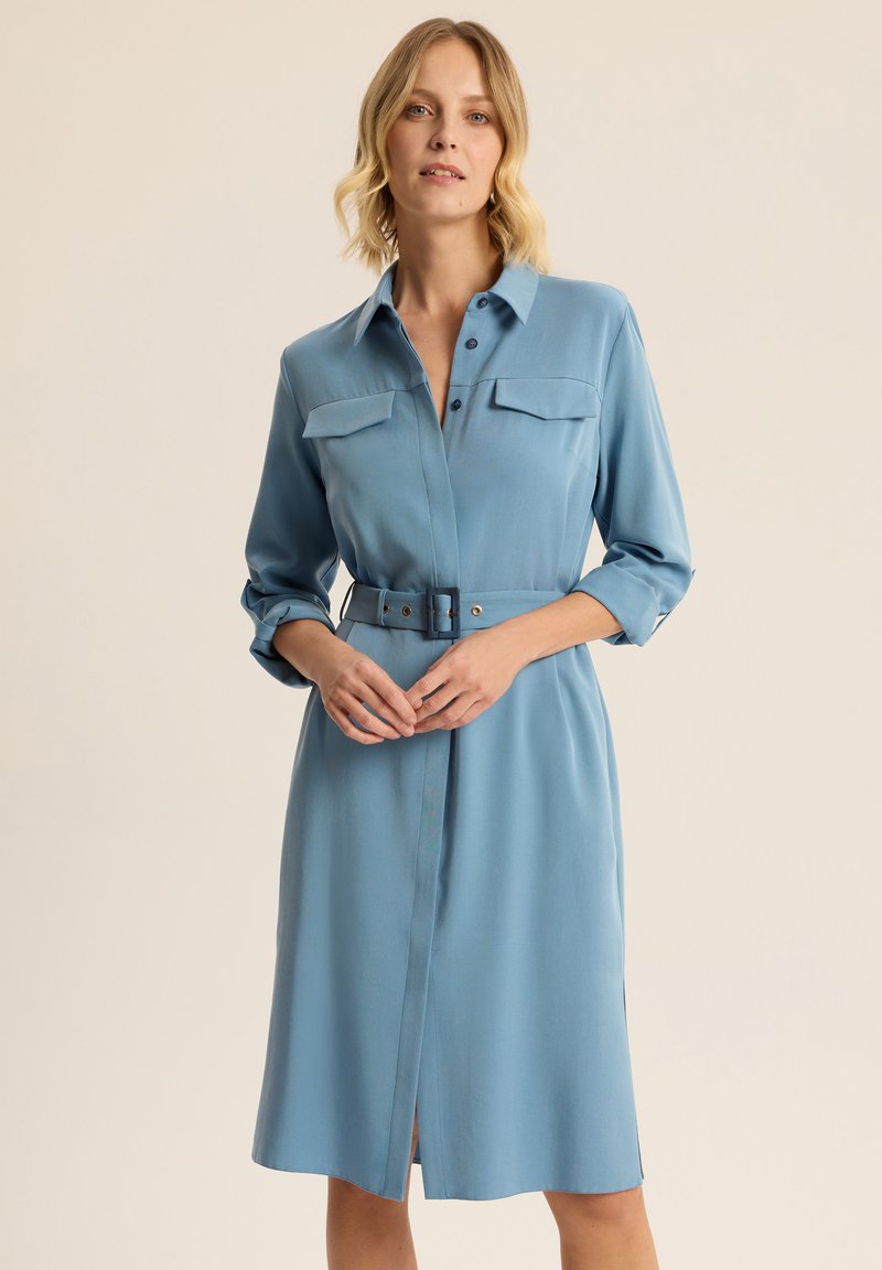 Niebieska sukienka koszulowa z gładkiego materiału, z paskiem w talii, rękawami o długości trzy czwarte oraz dwiema kieszeniami na piersi z guzikami.