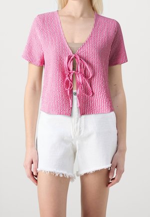 Mujer con una camiseta rosa texturizada de manga corta con lazos delanteros y shorts vaqueros blancos deshilachados sobre un fondo liso.