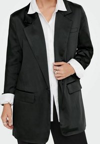 Blazer en satin noir avec une coupe ajustée, des revers crantés et deux poches avant, porté sur une chemise blanche à boutons.