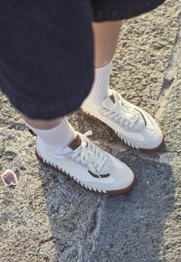 Bijela i siva tenisica s teksturiranim antilopom i kožom, s smešnim gumenim đonom, vezicama i oznakom brenda na jeziku.