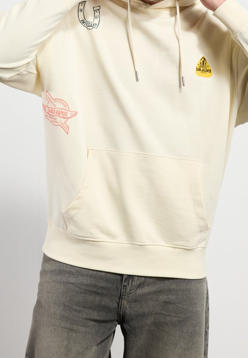 Personne portant un sweat à capuche crème avec poche avant et petits logos imprimés, associé à un jean gris délavé.